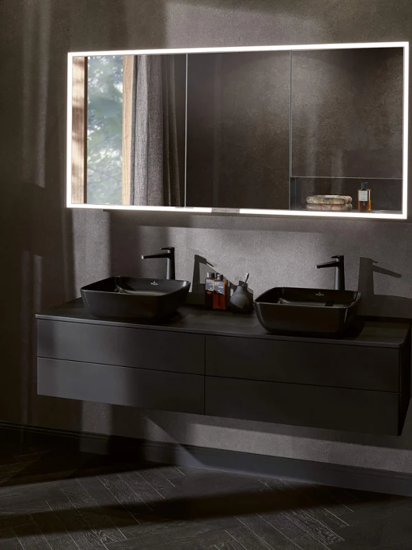 Lavabo Artis Pure Black dans une salle de bains noire Salle de bains moderne avec un meuble double vasque, un miroir rétroéclairé, un lampadaire rond et une serviette posée sur une petite table.