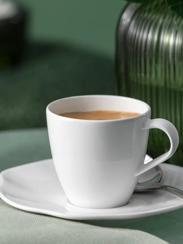 Une tasse à café Pura blanche avec soucoupe et cuillère est posée à côté d'un vase en verre vert. Une tasse à café Pura blanche avec soucoupe et cuillère est posée sur une surface verte à côté d'un vase en verre vert.