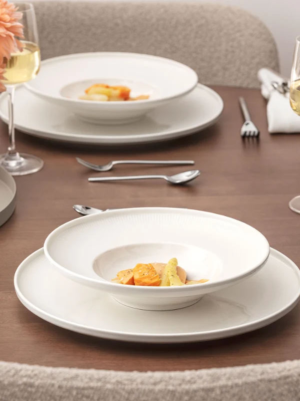 Une table bien dressée de la collection Afina avec deux assiettes garnies de plats gastronomiques, des verres à vin, des verres à eau, des couverts en argent, des serviettes en tissu et un vase avec des fleurs de pêcher sur une surface en bois.
