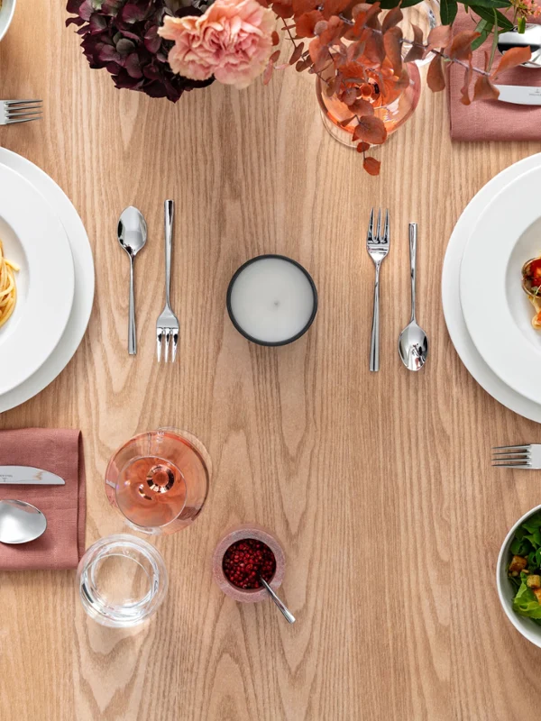 Table de salle à manger avec deux assiettes royales de pâtes, deux salades, des couverts, une bougie et une boisson. Table de salle à manger avec deux assiettes royales de pâtes, deux salades, des couverts, une bougie et une boisson. Les assiettes Villeroy & Boch sont posées sur des serviettes roses et une composition florale rose est disposée sur la table.