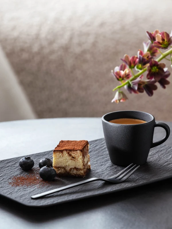Une tasse à expresso et une assiette rectangulaire Manufacture avec du gâteau Une part de tiramisu à côté d’une tasse à expresso Manufacture noire, garnie de myrtilles sur une assiette Manufacture rectangulaire moderne avec un vase décoratif et des fleurs en arrière-plan.