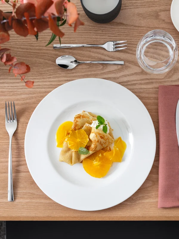 Crêpes aux oranges sur Royal Une table à manger élégamment dressée avec une assiette de crêpes garnies de tranches d'orange et de menthe. Les couverts sont placés à côté d'un verre et d'un bol contenant d'autres tranches d'orange.