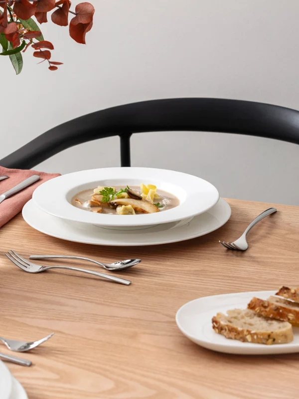 Table de salle à manger avec un bol de soupe et une assiette de la collection Royal de Villeroy & Boch Table de salle à manger avec un bol de soupe et une assiette de la collection Royal de Villeroy & Boch. Un verre d'eau et des couverts sont disposés sur une surface en bois clair. Chaise sombre et décoration végétale en arrière-plan.