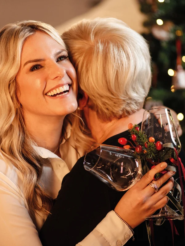 Deux femmes s’enlacent, l’une tient un verre à vin Toy’s Delight dans la main. Deux femmes s’enlacent devant un sapin de Noël décoré. La femme qui regarde l’appareil photo tient un verre à vin Toy’s Delight.