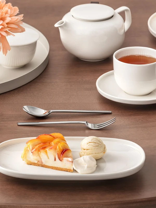 La table dressée de la collection Afina présente une assiette avec un gâteau et une glace, une tasse de thé et une théière posées sur une table en bois avec un vase à fleurs.
