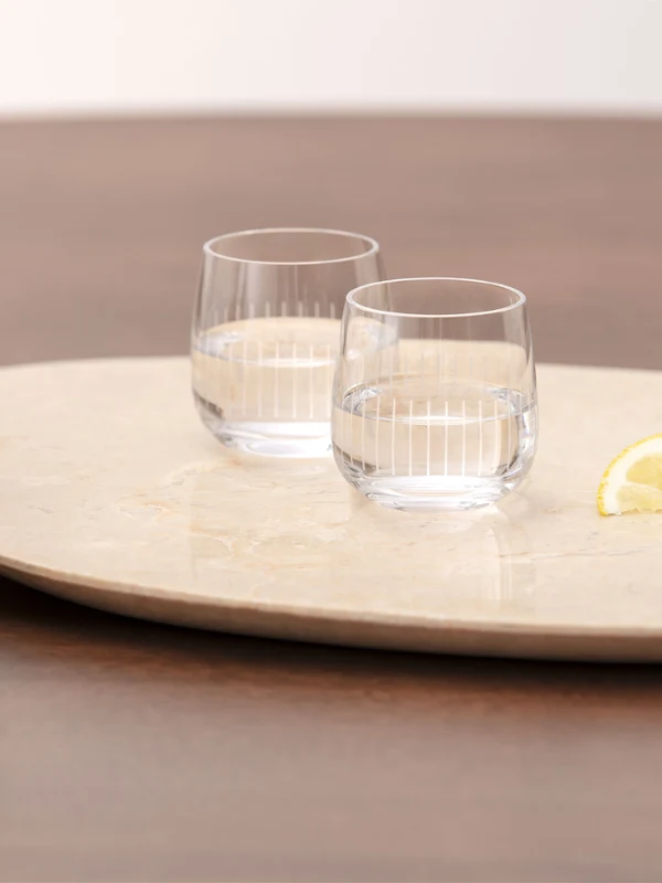 Deux verres à eau transparents de la collection Afina, deux quartiers de citron et un bol de noix sont posés sur un plateau rond posé sur une table en bois. À l'arrière-plan, on aperçoit un vase contenant une fleur couleur pêche.