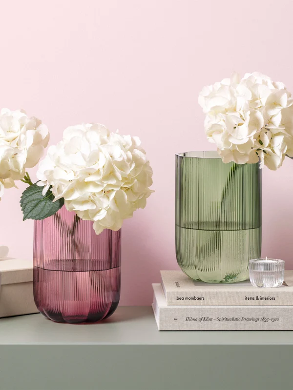 Deux vases en verre Fleur avec des hortensias blancs sur une table à côté de livres empilés, d'un coffret cadeau et d'une petite lampe verte.