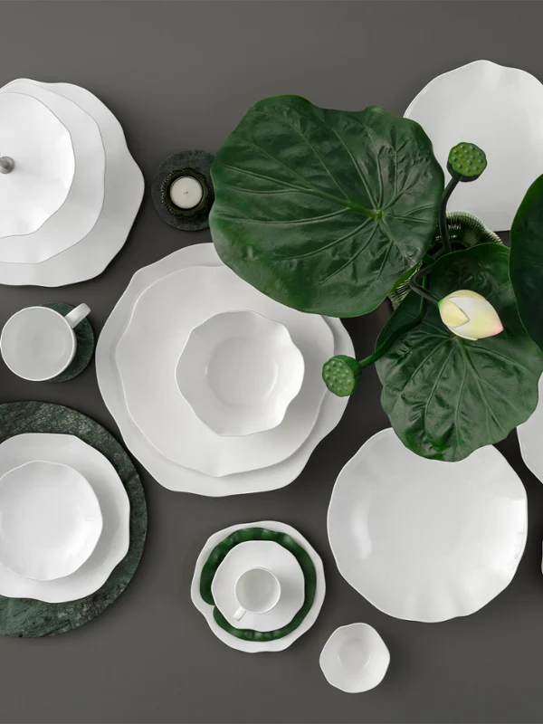 Des pièces de vaisselle blanches de la collection Pura posées sur une surface sombre. Vaisselle blanche de la collection Pura sur un fond sombre décoré de grandes feuilles vertes et d'une seule fleur blanche.