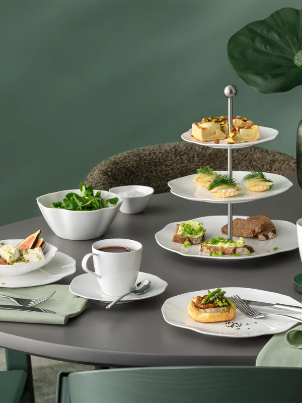 Une table ronde avec de la vaisselle blanche de la collection Pura devant un fond vert Une table ronde avec de la vaisselle blanche de la collection Pura, divers produits de boulangerie, de la salade, du thé et un décor de feuilles vertes sur fond vert.