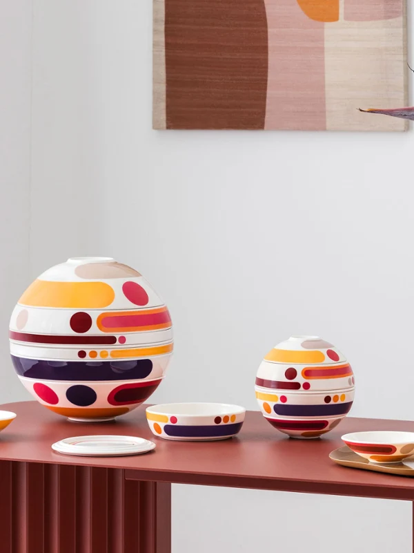 La petite boule miami et la boule miami disposées sur une table rouge Des services de table colorés et composés de plusieurs pièces de la petite boule miami, objets design circulaires de grande et petite taille, sont disposés à titre décoratif à côté d'un vase aux motifs abstraits orné de fleurs orange sur une table rouge.