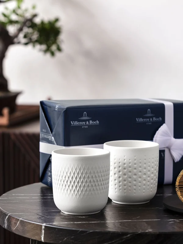 Deux tasses de collier blanches devant un bonsaï Deux tasses à collier blanches sur une table avec un fouet à thé et un paquet cadeau, devant un bonsaï.