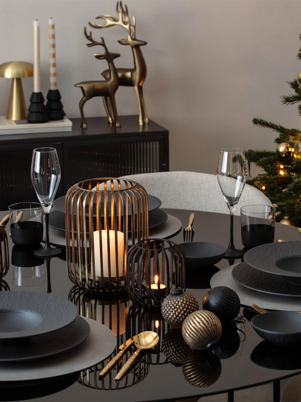 A black dining table set with dark crockery Une table à manger noire dressée avec de la vaisselle foncée, des décorations de Noël, des bougies et un sapin de Noël illuminé en arrière-plan.