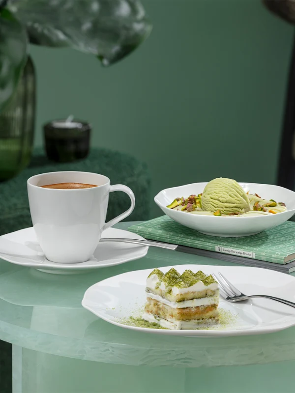 Une tasse à café Pura, une part de gâteau et une coupe à dessert Pura sont posées sur une table en verre. Une tasse à café Pura, une part de gâteau et une coupe à dessert Pura avec de la glace sont posées sur une table en verre avec une plante verte en arrière-plan.