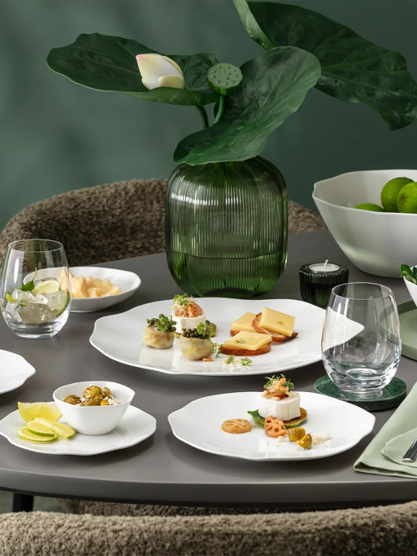 Une table ronde dressée avec la vaisselle blanche de la collection Pura. Une table ronde dressée avec la vaisselle blanche de la collection Pura, comprenant des assiettes à entrée, des verres, des serviettes et un centre de table composé de plantes vertes.