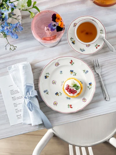 Une assiette Petite Fleur avec une tarte, une tasse à thé Une assiette Petite Fleur avec un gâteau, une tasse à thé, une boisson avec une fleur et une serviette sur une table en bois.