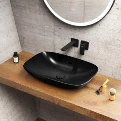 Une salle de bains moderne avec une vasque à poser noire Loop & Friends de Villeroy & Boch sur un plan de toilette en bois, un robinet mural noir, un miroir rond et des articles de toilette comprenant du savon, un rasoir, une brosse et une brosse à dents dans un support.