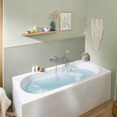 Une baignoire O.novo remplie d’eau Une salle de bains moderne avec une baignoire remplie d’eau de la collection O.novo, une étagère murale avec des articles de toilette, une tenture murale en macramé et une fenêtre avec vue sur la verdure.
