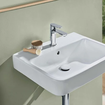 Un lavabo blanc O.novo avec une robinetterie chromée Un lavabo moderne rectangulaire blanc O.novo avec un robinet chromé, une brosse et un récipient devant un mur vert mat. Une chaise beige se trouve à l’arrière-plan.