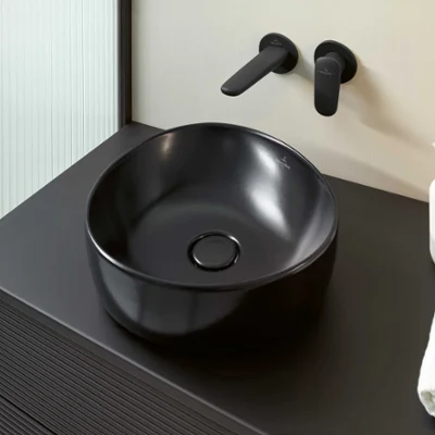 Un ensemble de salle de bains moderne avec une vasque ronde noire à poser Antao de Villeroy & Boch, des robinets noirs montés au mur, un distributeur de savon liquide blanc et une serviette blanche pliée sur un plan de toilette noir.