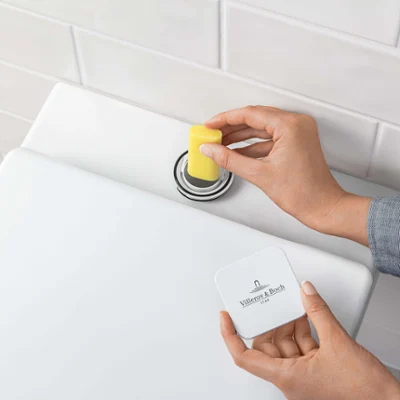 Une personne jette un bloc WC jaune dans le réservoir de chasse d’eau de toilettes. En arrière-plan, on peut voir des murs carrelés en blanc.