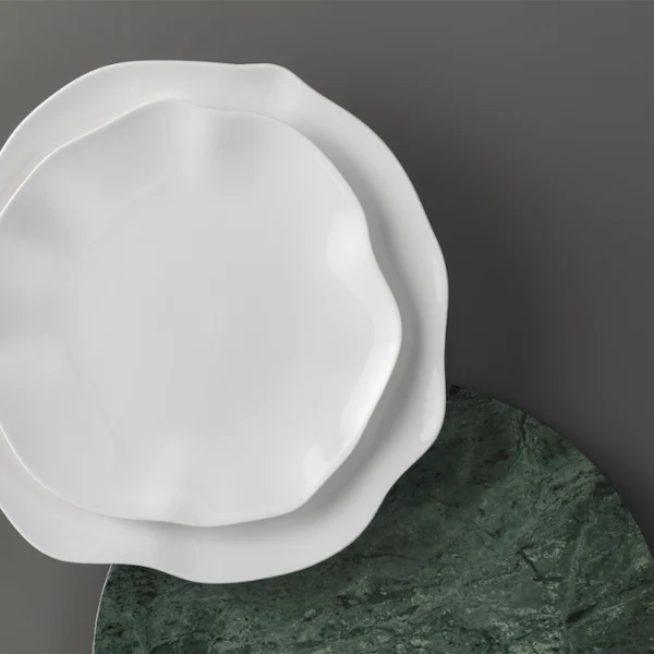 Deux assiettes Pura blanches de forme irrégulière sont empilées sur un fond gris. Deux assiettes Pura blanches de forme irrégulière sont empilées sur une assiette ronde vert foncé devant un fond gris.