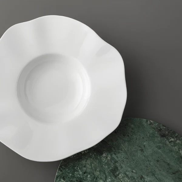 Une assiette Pura blanche avec un bord festonné sur une surface grise Une assiette Pura blanche à bord festonné est placée sur une surface grise à côté d'un tapis rond vert texturé.