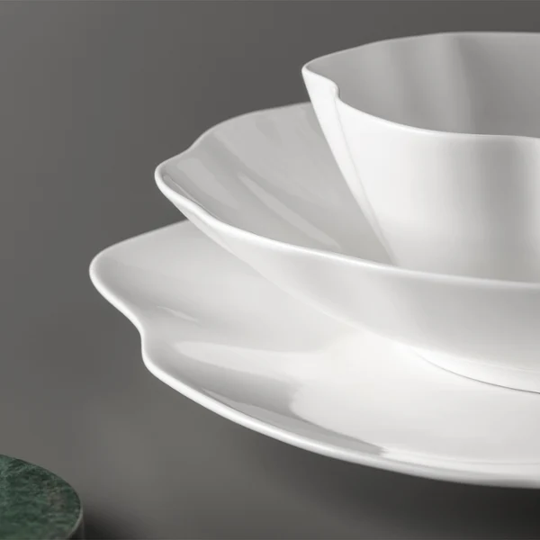 Trois assiettes blanches Pura à bord dentelé sont posées sur une surface grise. Trois assiettes blanches Pura à bord dentelé sont empilées sur une surface grise à côté d'un objet rond vert.