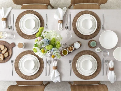 Table Artesano vue du ciel Table de salle à manger pour six personnes dressée avec de la porcelaine de Villeroy & Boch, des sets de table bruns, des serviettes blanches et une composition florale. Du pain, des pâtes à tartiner et des épices sont également disponibles.