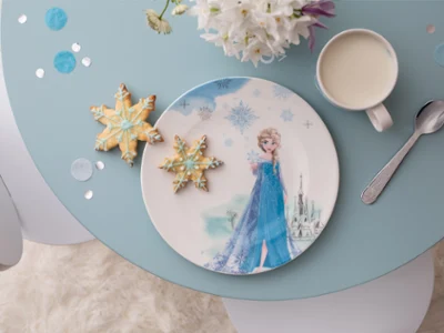 Une assiette avec une figurine Frozen Une assiette avec une figurine Frozen, deux biscuits en forme de flocon de neige, une tasse de lait et une cuillère sur une table bleue.