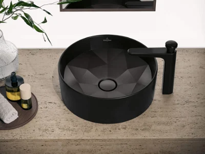 Lavabo Octagon noir sur un plan de toilette en béton Lavabo rond noir mat avec robinet assorti sur un plan de travail moucheté ; miroir et plante à proximité.