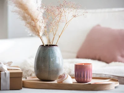 Un plateau en bois avec un vase Lave et une bougie Perlemor Un plateau en bois sur lequel sont posés un vase Lave et une bougie Perlemor avec des fleurs séchées, à côté d’un cadeau emballé.