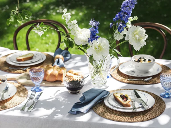 Une table dressée avec de la vaisselle Vieux Luxembourg dans un jardin Une table dressée en plein air avec des fleurs, des verres bleus, du pain et des assiettes Vieux Luxembourg contenant de la nourriture sur des sets de table tissés dans un environnement de jardin.