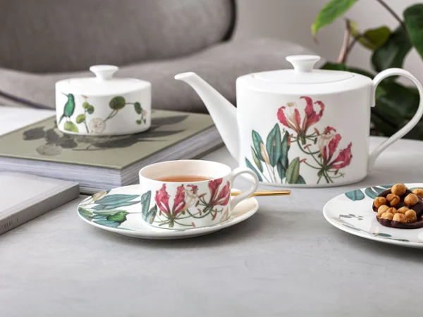 Une théière, une tasse et une assiette avec des snacks au motif floral sont disposées sur une table à côté de livres et d'une plante en arrière-plan.