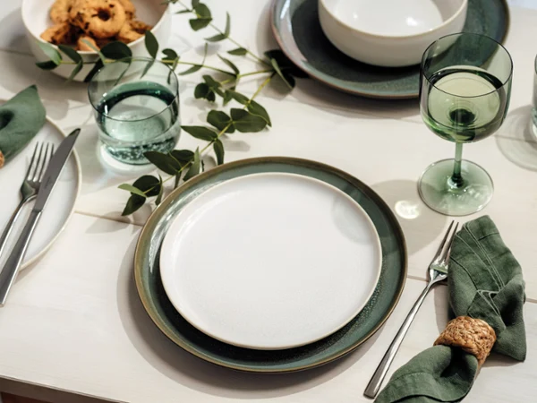 Une table de salle à manger joliment dressée avec des assiettes vertes, des verres, des bols, des serviettes et des plantes vertes en guise de décoration.