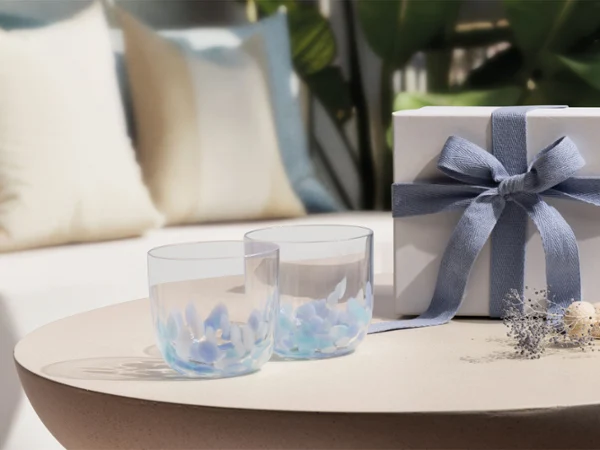 Deux verres transparents Like Aqua et un coffret cadeau blanc Deux verres transparents Like Aqua et un coffret cadeau blanc avec un ruban bleu sur une table ronde ; canapé et coussins en arrière-plan.