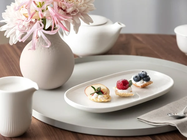 Petites douceurs sucrées avec Afina de Villeroy & Boch Une assiette blanche Afina avec trois petites pâtisseries et des fruits se trouve sur un plateau rond à côté d'un vase à fleurs, d'une théière et d'un pot à lait sur une table en bois.