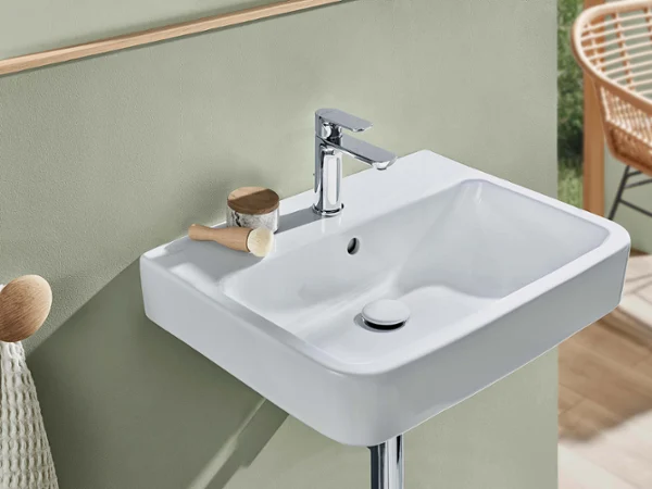 Un lavabo blanc O.novo avec une robinetterie chromée Un lavabo moderne rectangulaire blanc O.novo avec un robinet chromé, une brosse et un récipient devant un mur vert mat. Une chaise beige se trouve à l’arrière-plan.