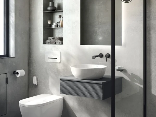 Une salle de bains moderne avec des murs gris, un plan de toilette suspendu, un lavabo blanc, une robinetterie murale et un miroir rétroéclairé. Des toilettes et étagères ouvertes avec des serviettes et des articles de toilette. Une cabine de douche en verre visible à droite.