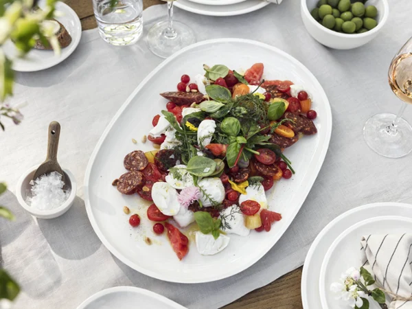 Sur une nappe blanche se trouve un plateau Artesano de Villeroy & Boch avec une salade mixte composée de tomates, d’herbes aromatiques et de charcuterie. À côté, se trouvent des coupes contenant des olives, un verre de rosé et une coupe de sel.