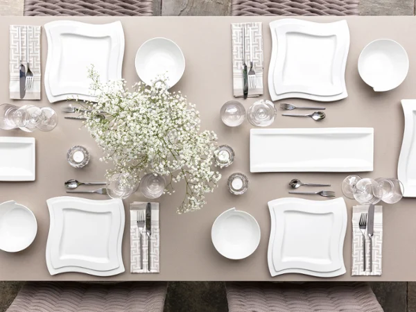 Vue de dessus d'une élégante table de salle à manger pour cinq personnes, avec des assiettes carrées blanches NewWave. Vue de dessus d'une élégante table de salle à manger pour cinq personnes, avec des assiettes carrées blanches NewWave de Villeroy & Boch, des serviettes à motifs, des verres, des couverts et un centre de table floral avec de petites fleurs blanches.
