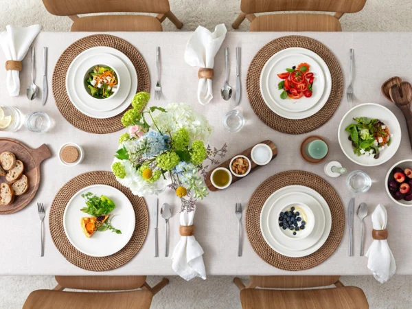 Vue de dessus d'une table à manger joliment dressée avec des salades, des fruits, du pain, des verres d'eau et des fleurs au centre sur une nappe beige.