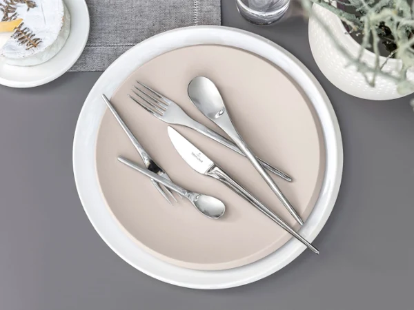 Des couverts de table NewMoon sur une assiette beige Des couverts de table NewMoon de Villeroy & Boch disposés sur une assiette beige placée sur un fond gris.
