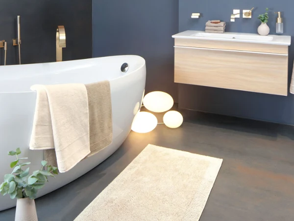 Une baignoire Villeroy & Boch blanche avec des meubles de salle de bains en bois Une salle de bains moderne avec une baignoire blanche en îlot, un lavabo suspendu, des robinetteries dorées, des serviettes, une plante en pot et des boules lumineuses sur le sol