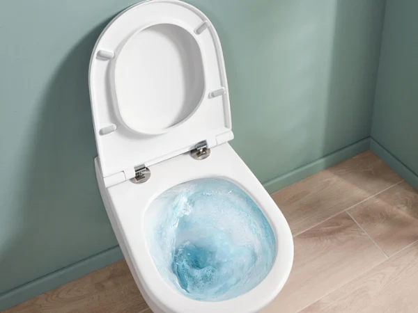 Une cuvette de toilettes blanche ouverte avec de l’eau de chasse d’eau bleue, devant un mur bleu-vert et un sol en bois clair.