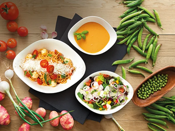 Un ensemble starter Vapiano de Villeroy & Boch rempli de nourriture Un ensemble de base Vapiano de Villeroy & Boch sur une table en bois avec des tomates, des oignons nouveaux, des petits pois, des haricots et des oignons. Des assiettes Vapiano garnies de pâtes, de la salade et de la soupe de potiron