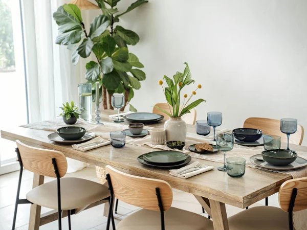 Le set de table couvert avec différents éléments de la collection Lave de Villeroy & Boch Le set de table pour la salle à manger, avec différents éléments de la collection Lave de Villeroy & Boch en vert et gris, accentués par des verres et des serviettes bleus. Un vase avec des fleurs jaunes et une petite plante en pot assurent la décoration, tandis qu'une grande plante est placée près de la fenêtre.