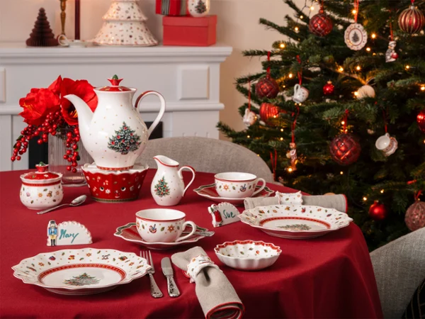 A table set with crockery and cutlery Une table dressée avec de la vaisselle et des couverts aux motifs de Noël, à côté d'un sapin de Noël décoré et d'une cheminée illuminée.
