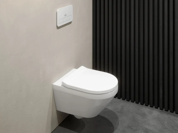 Des toilettes Architectura de Villeroy & Boch en blanc Des toilettes Architectura blanches modernes à couvercle fermé, suspendues, côtoient un mur beige et un panneau noir texturé. Les toilettes sont équipées d’une chasse d’eau minimaliste à deux débits.