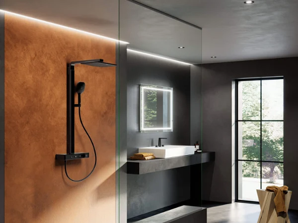 Une salle de bains moderne au design minimaliste avec une douche vitrée, une pomme de douche noire fixée au mur, un grand miroir avec éclairage, un lavabo avec plan de toilette sombre et un tabouret près d’une fenêtre.