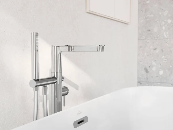 Une robinetterie de baignoire moderne chromée avec douchette à main est installée devant un mur carrelé blanc. La baignoire blanche est partiellement visible au premier plan.